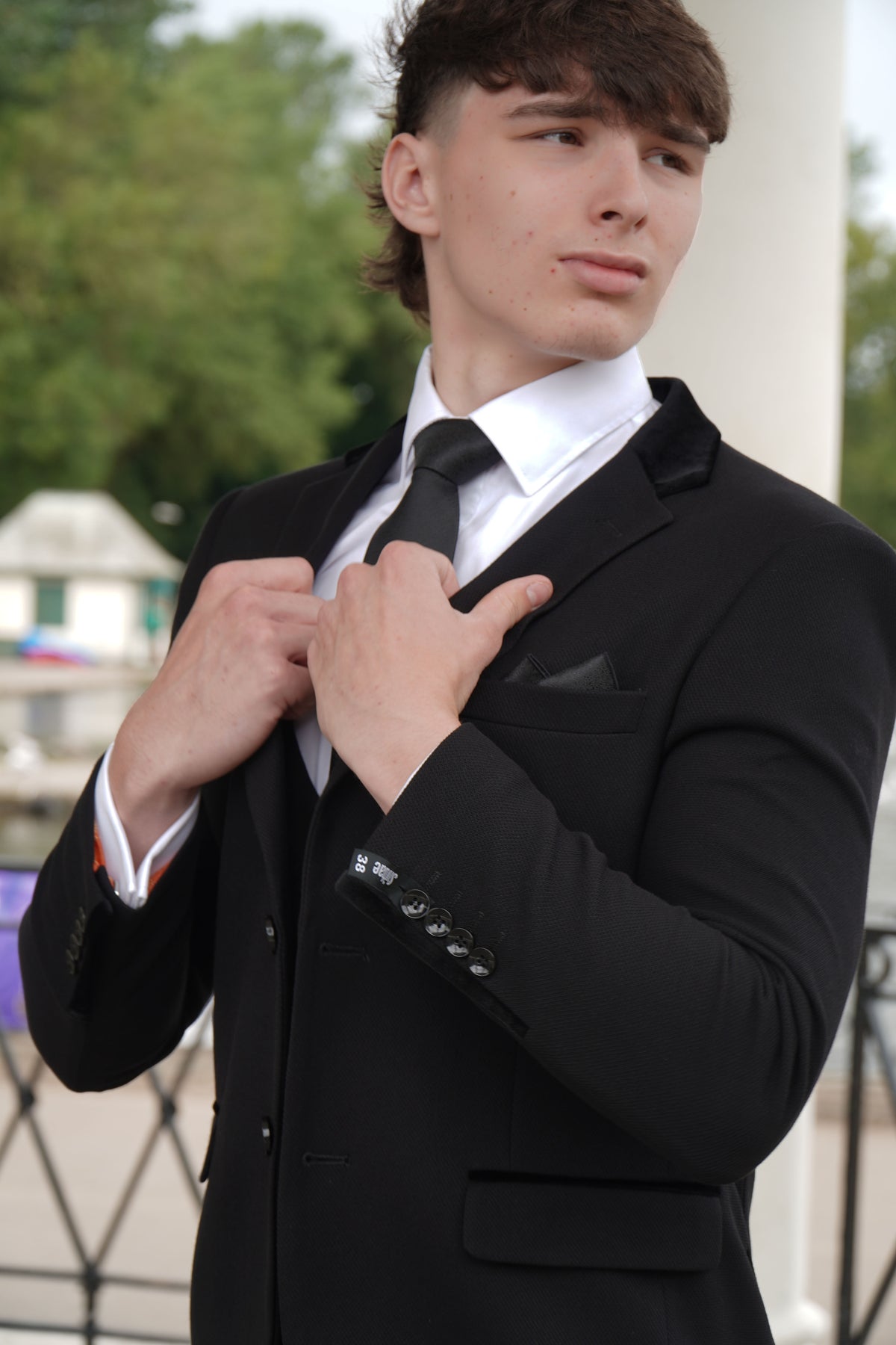 A man modeling a Suitbae black 3 piece suit with velvet lapel accents, a white dress shirt, and a black satin tie outdoors.