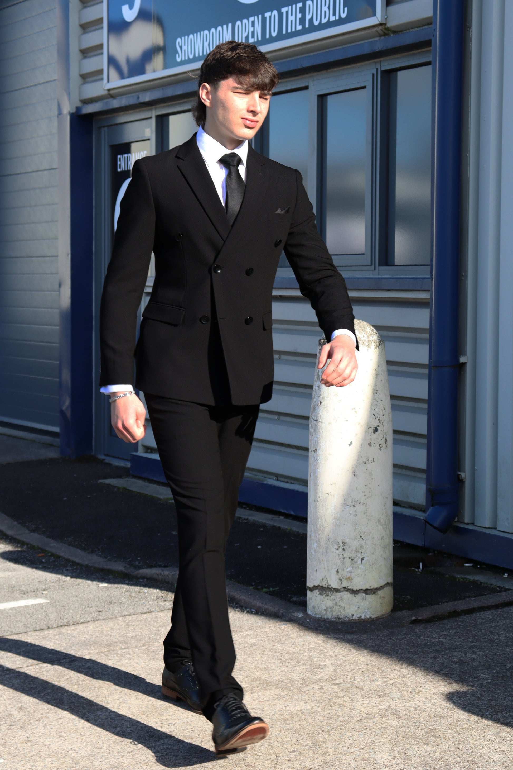 Man walking confidently outside the SUITBAE store wearing the Diablo Black Double Breasted Three-Piece Suit, paired with a white shirt, black tie, and black brogues for a sleek formal look.
