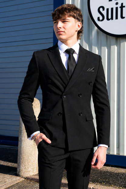 A young man wearing a black double-breasted suit, white dress shirt, and black tie, standing outdoors in front of a blue and white building.