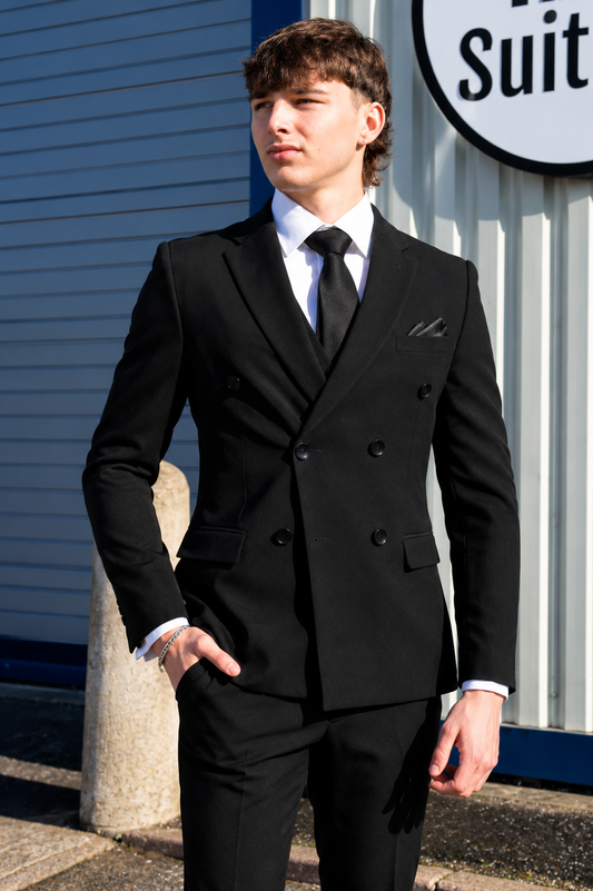 A young man wearing a black double-breasted suit, white dress shirt, and black tie, standing outdoors in front of a blue and white building.