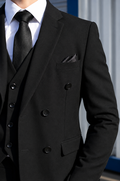 A close-up of a man wearing a black double-breasted suit jacket with a matching vest and tie, paired with a white dress shirt and a black pocket square.