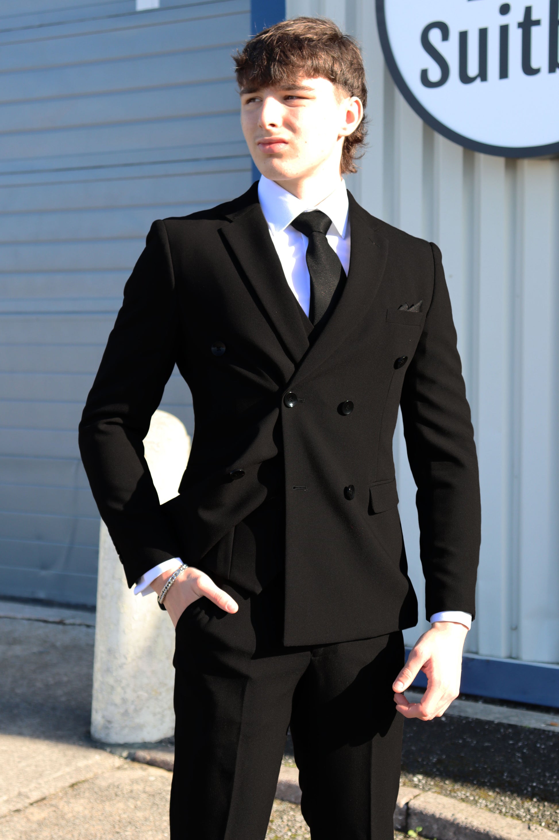 Man wearing the SUITBAE Diablo Black Double Breasted Three-Piece Suit, featuring peak lapels and a sharp tailored fit, styled with a white shirt and black tie outside the SUITBAE store.